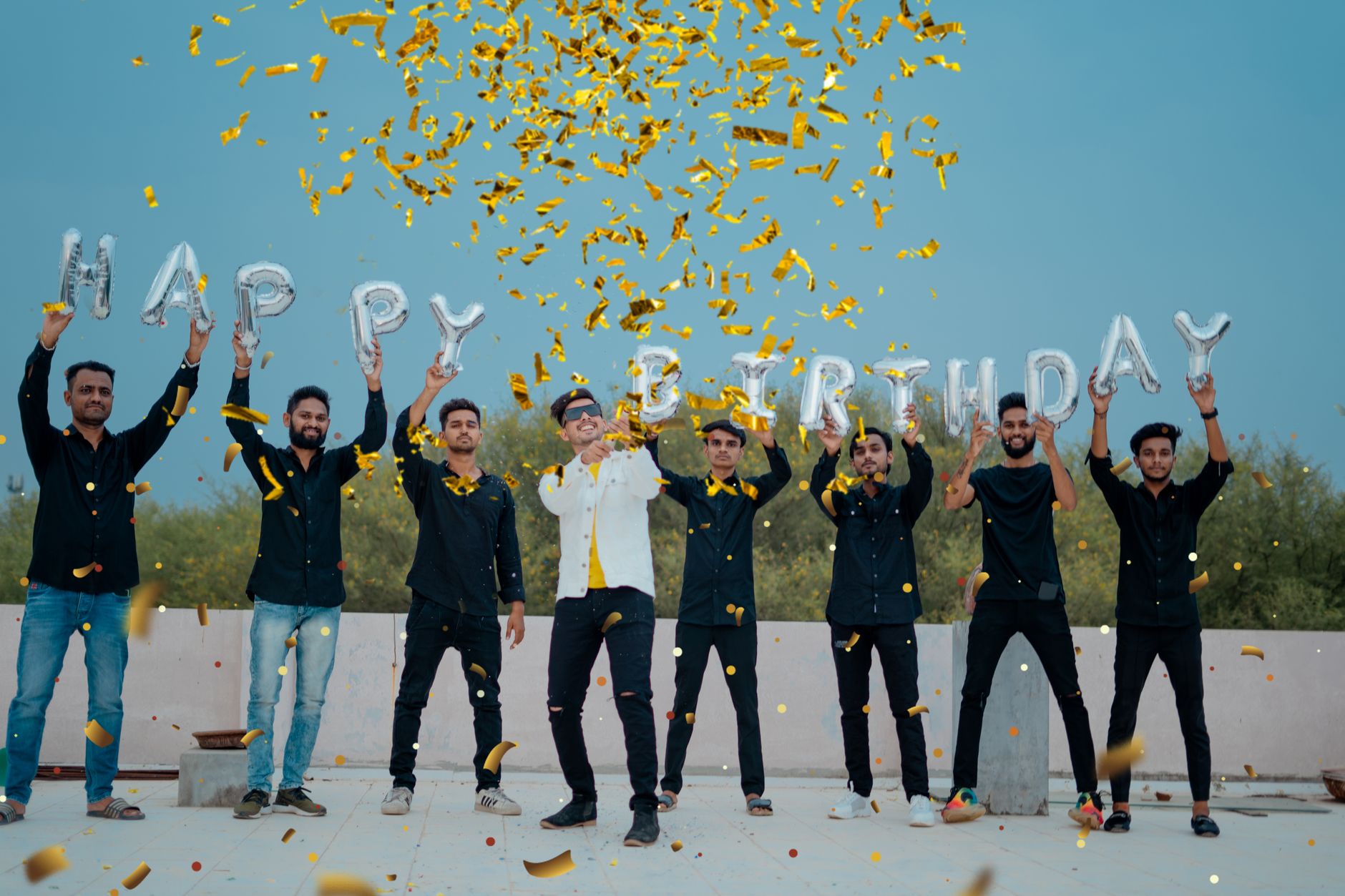 A joyful group of young men celebrates a birthday outdoors with confetti and balloon letters.