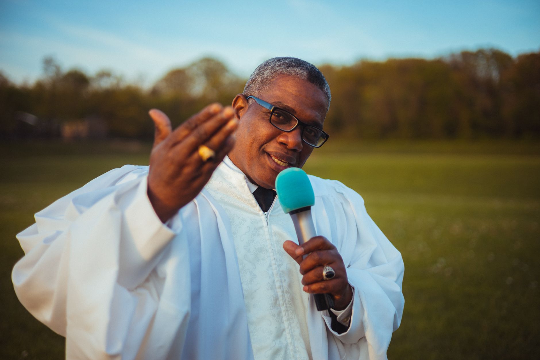 A priest gestures while speaking with a microphone in a meadow setting.