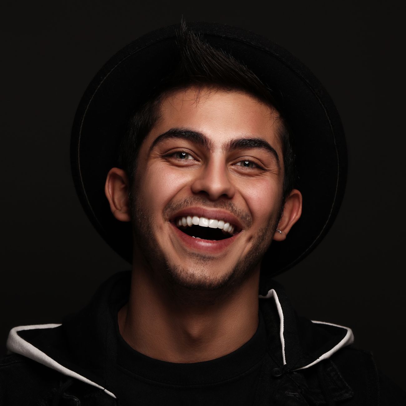 A joyful portrait of a young man smiling broadly, wearing a stylish black hat.